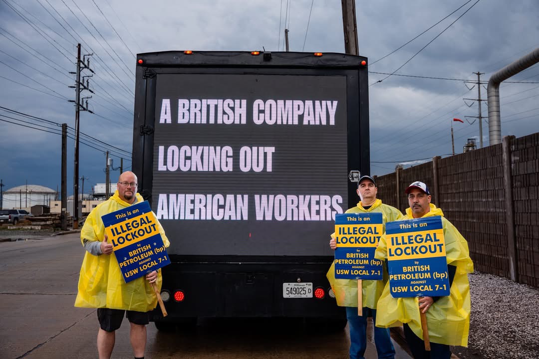 Three USW workers in rain ponchos holding illegal lockout signs in front of the AOTC LED truck